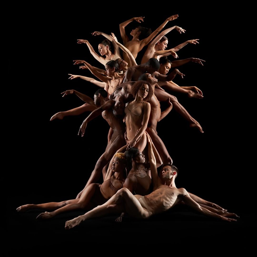 Striking studio portrait of a large group of nude male and female dancers arranged in a pyramid formation against a solid black background.