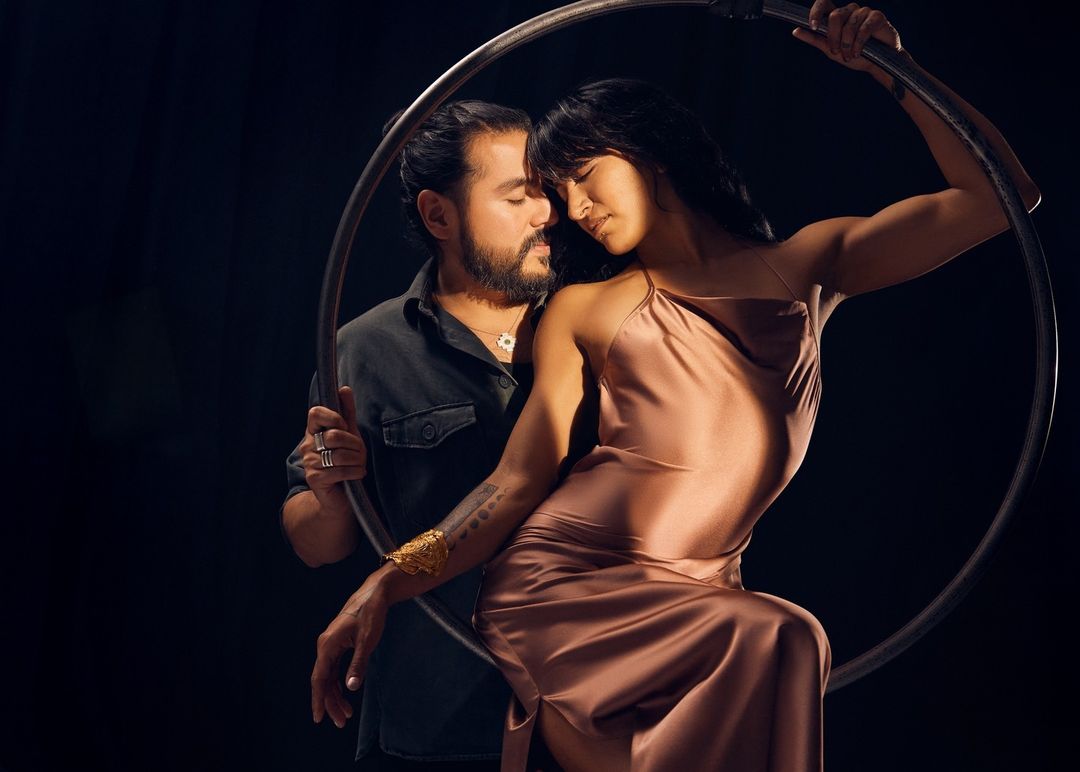 Artistic image of a couple posing together, framed within a circular metal hoop, set against a dark backdrop with soft lighting.