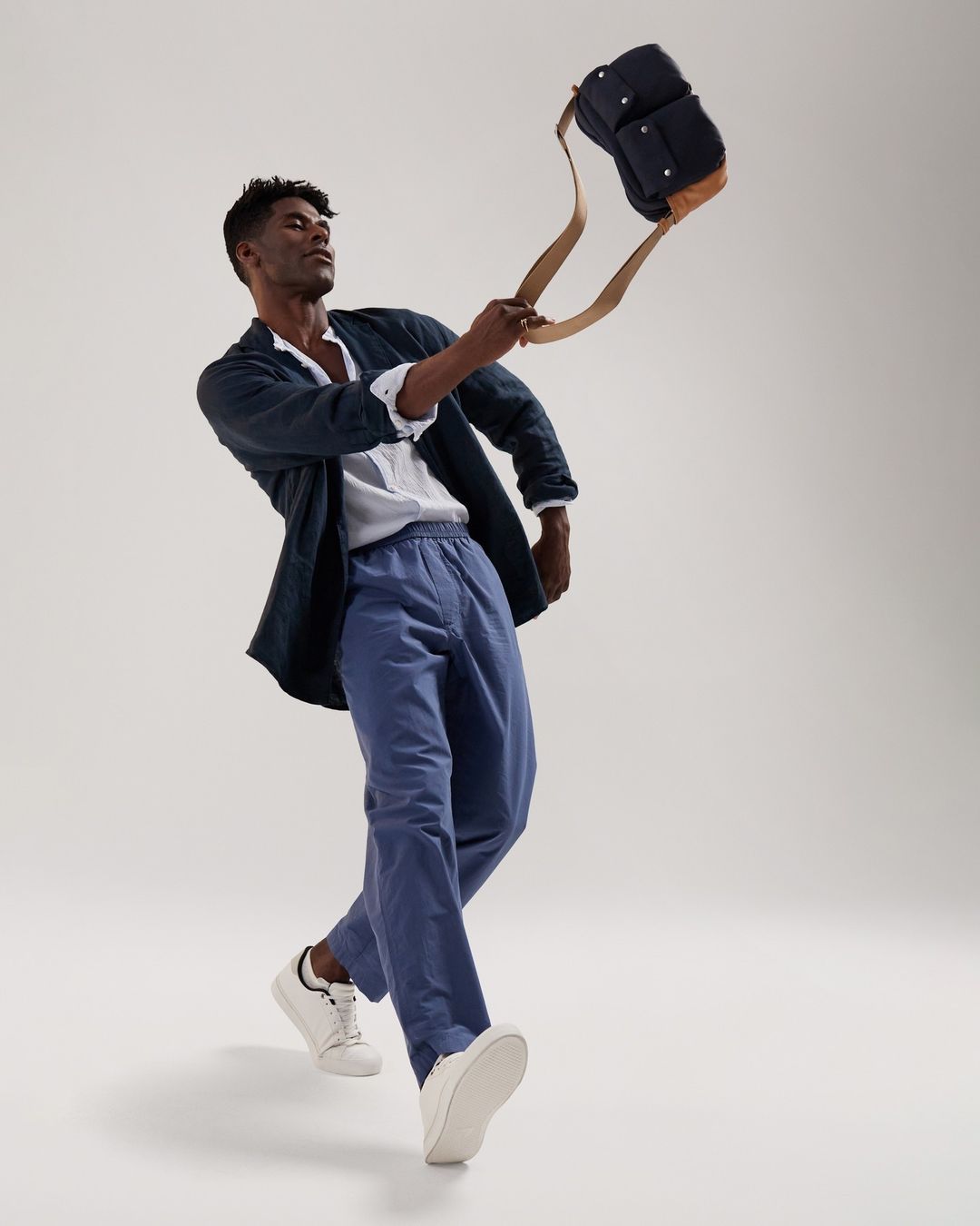 A man in casual clothing, including blue pants, a white shirt, and a dark jacket, holding a dark blue bag with tan straps in a studio setting.