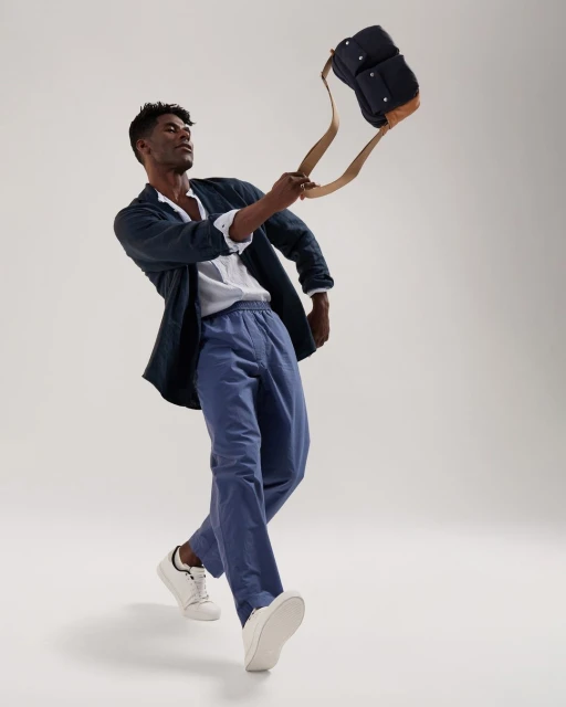 A man in casual clothing, including blue pants, a white shirt, and a dark jacket, holding a dark blue bag with tan straps in a studio setting.
