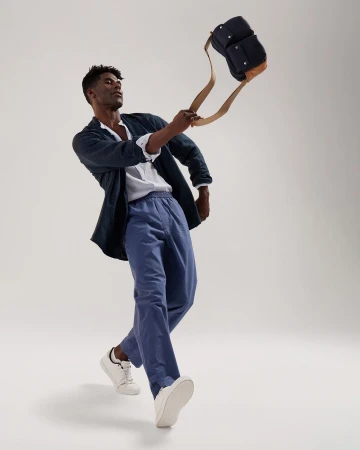 A man in casual clothing, including blue pants, a white shirt, and a dark jacket, holding a dark blue bag with tan straps in a studio setting.