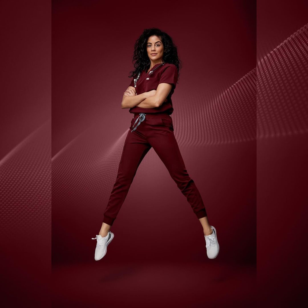Full-length shot of a woman in burgundy scrubs mid-air with arms crossed and legs splayed on a burgundy background with woven design.