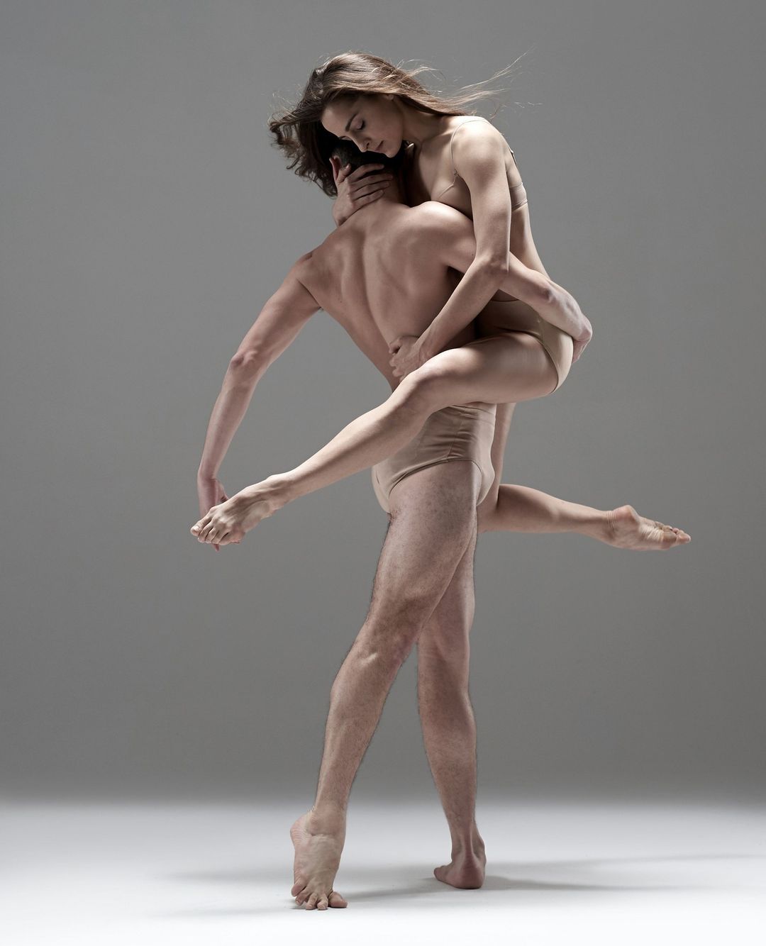 A male dancer supports a female dancer in a studio. They are both wearing body suits, and the lighting is diffused and even.