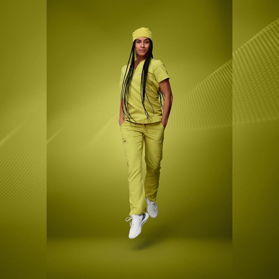 Full shot of a young adult woman with dark hair wearing yellow scrubs and white shoes, posing against a yellow background with abstract design.
