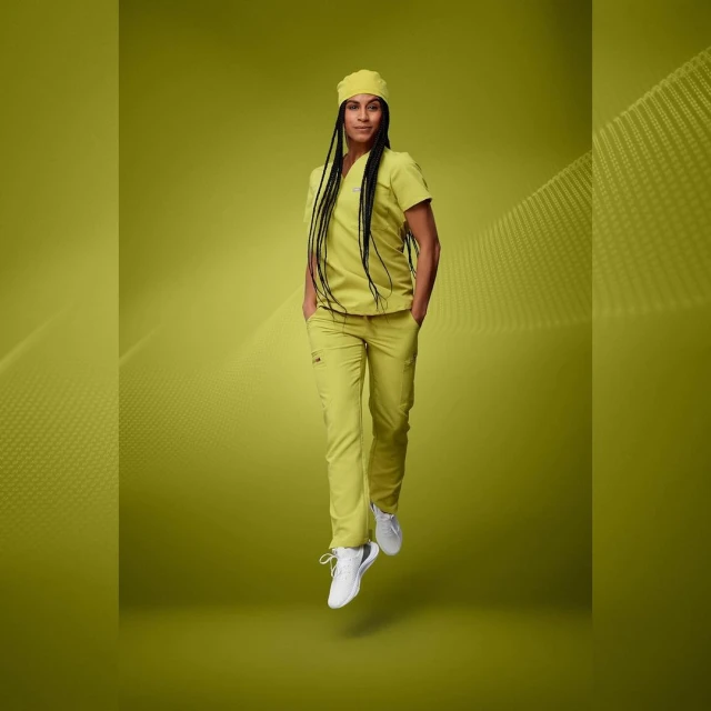 Full shot of a young adult woman with dark hair wearing yellow scrubs and white shoes, posing against a yellow background with abstract design.