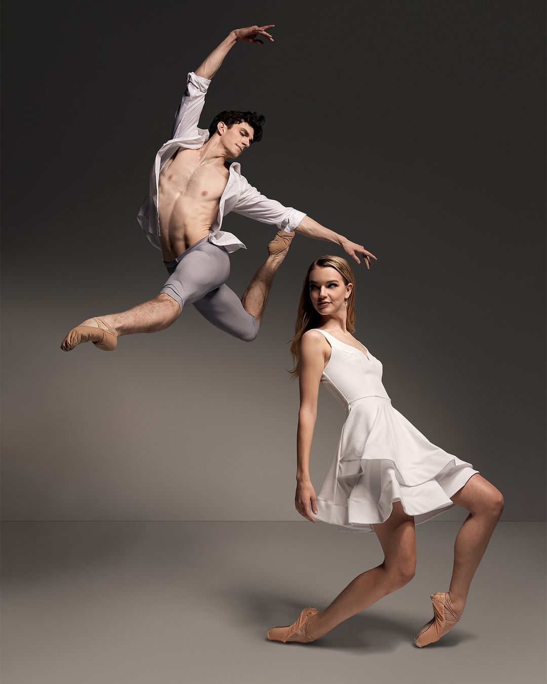 Male ballet dancer mid-air leap near ballerina in white dress on grey background, performing a dance routine in a studio.