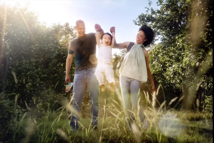 A happy family of three, including a child, is swinging outside in a grassy field surrounded by trees with warm sunlight.