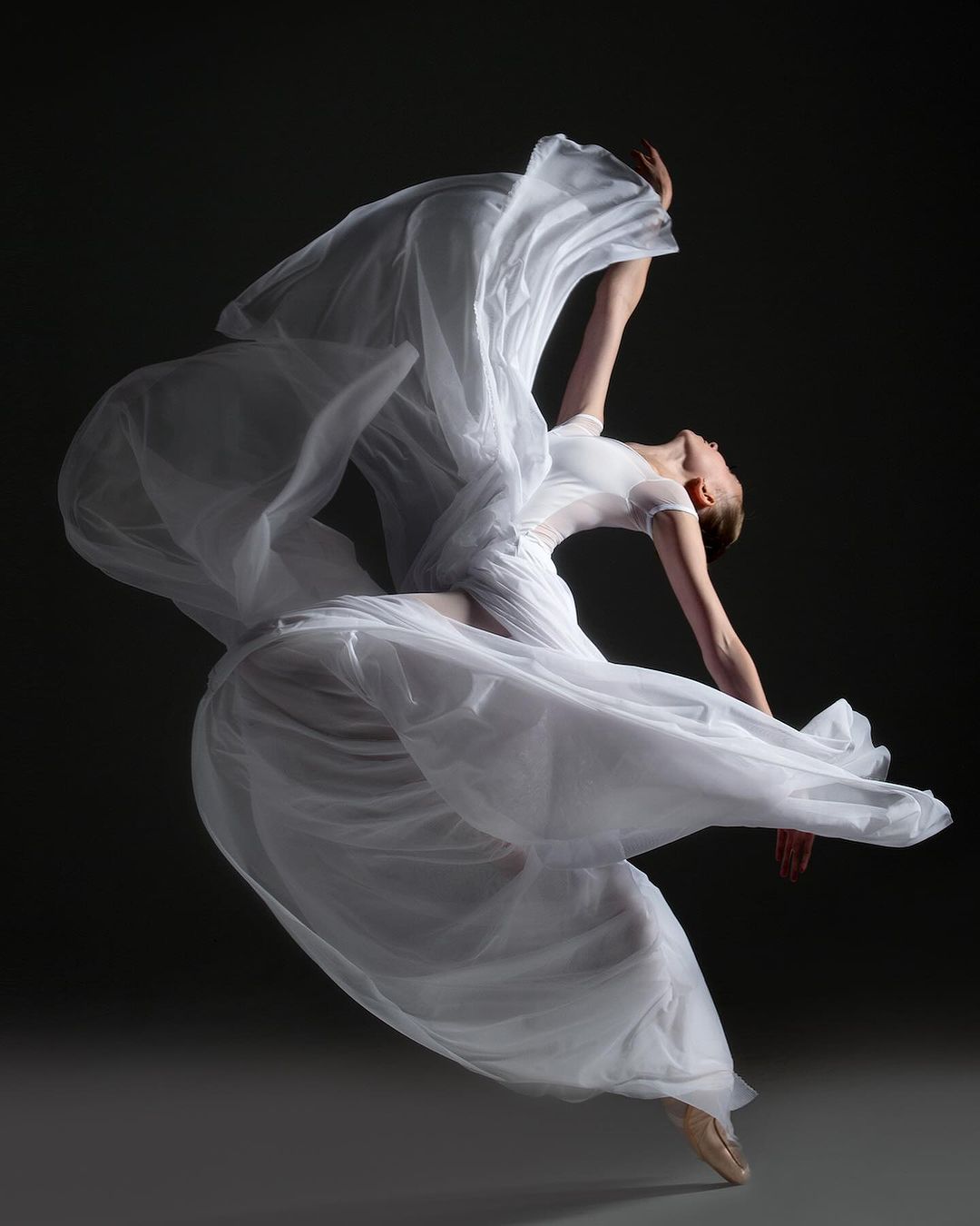 Elegant female ballet dancer in flowing white dress leaps and arches backward in a graceful pose against a dark gray background.