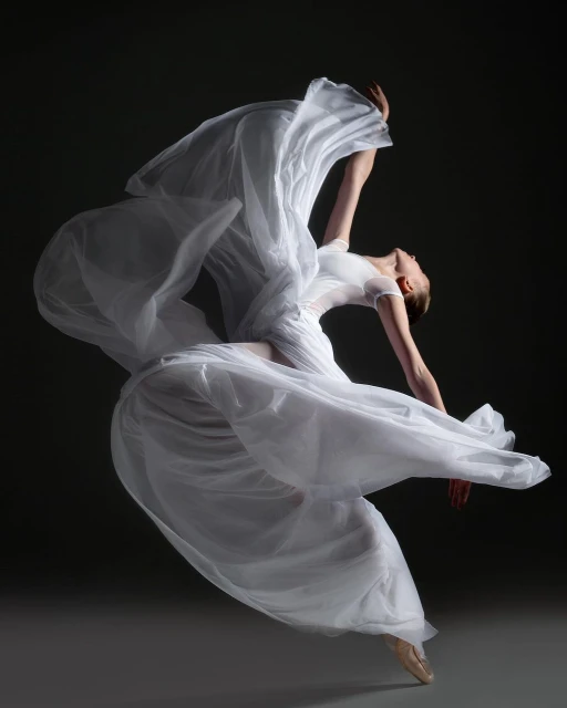Elegant female ballet dancer in flowing white dress leaps and arches backward in a graceful pose against a dark gray background.