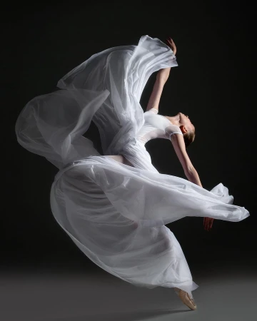 Elegant female ballet dancer in flowing white dress leaps and arches backward in a graceful pose against a dark gray background.