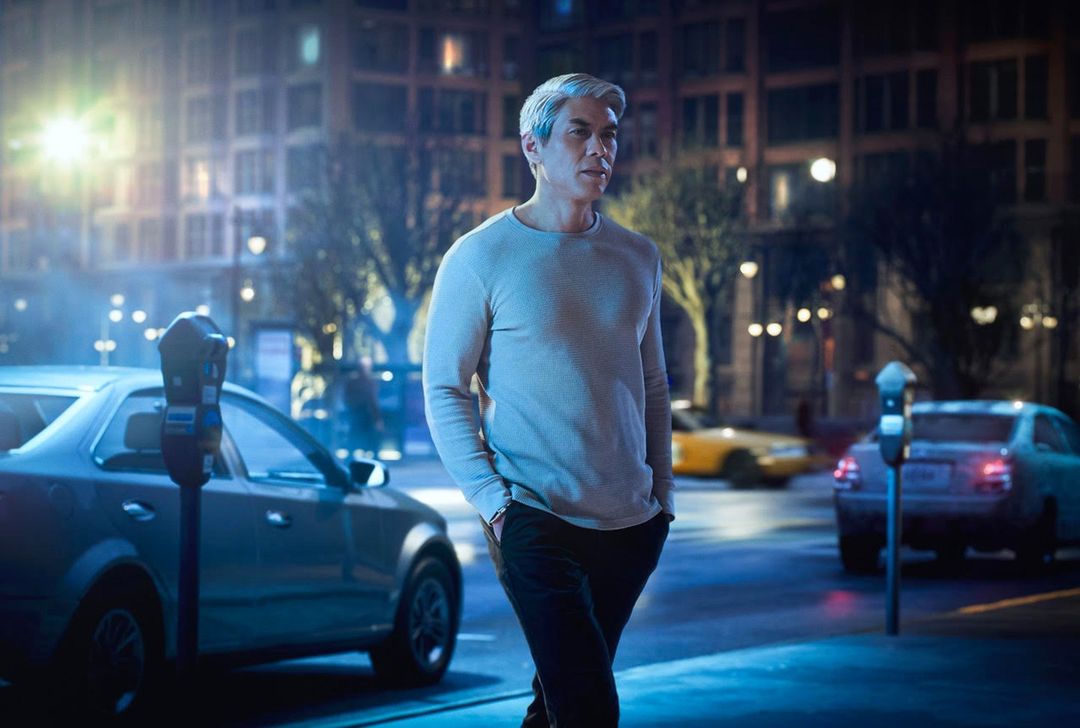 An adult male with graying hair walks on a city street at night, wearing a long-sleeved shirt and dark pants, with cars and buildings in background.