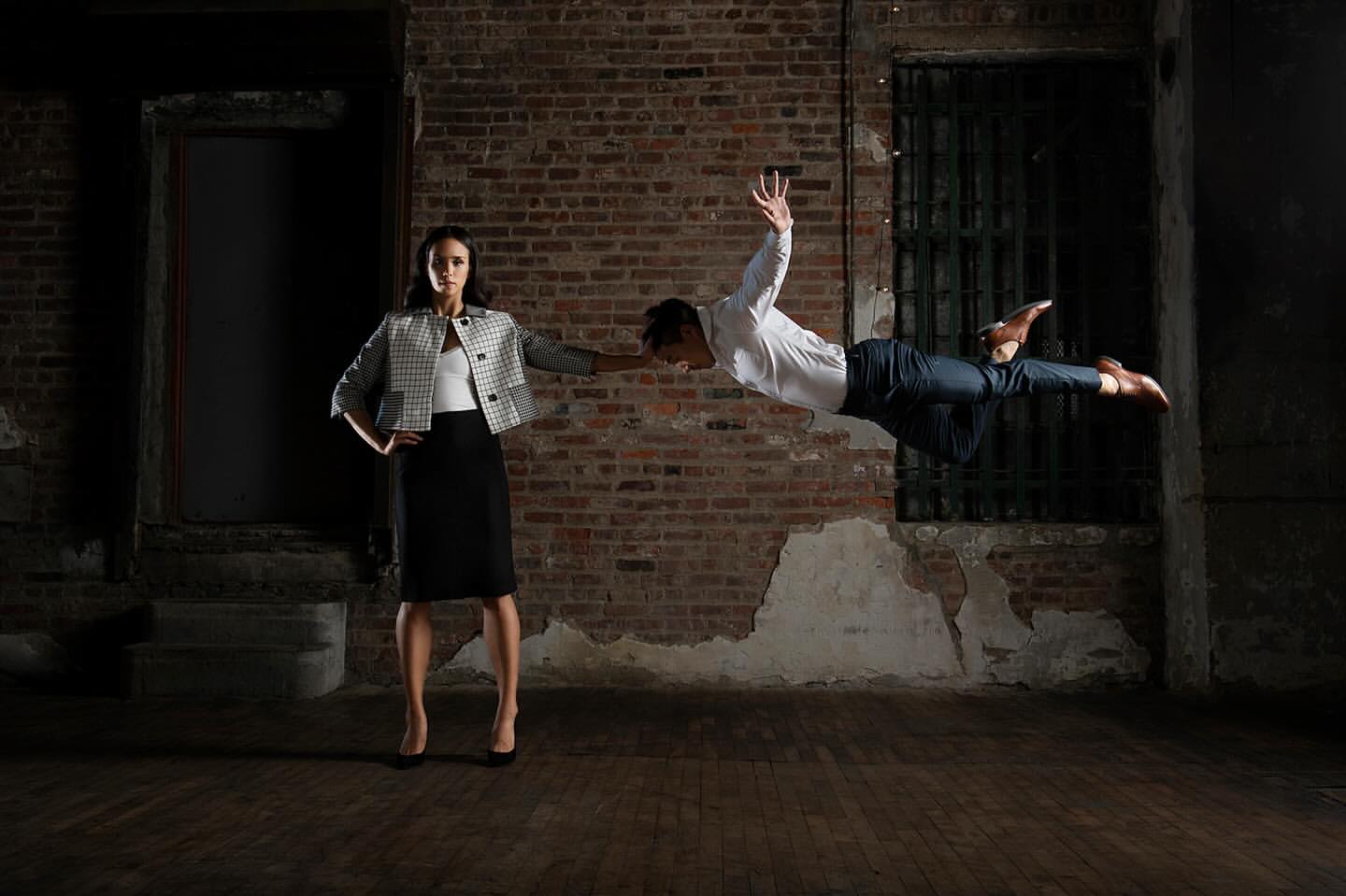 A woman standing with one hand extended while a man floats horizontally in mid-air beside her against an exposed brick wall.