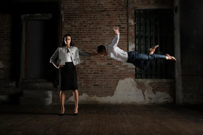 A woman standing with one hand extended while a man floats horizontally in mid-air beside her against an exposed brick wall.