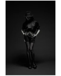 Model wearing a fedora hat, black blouse, shorts, and boots standing against a dark seamless studio background.