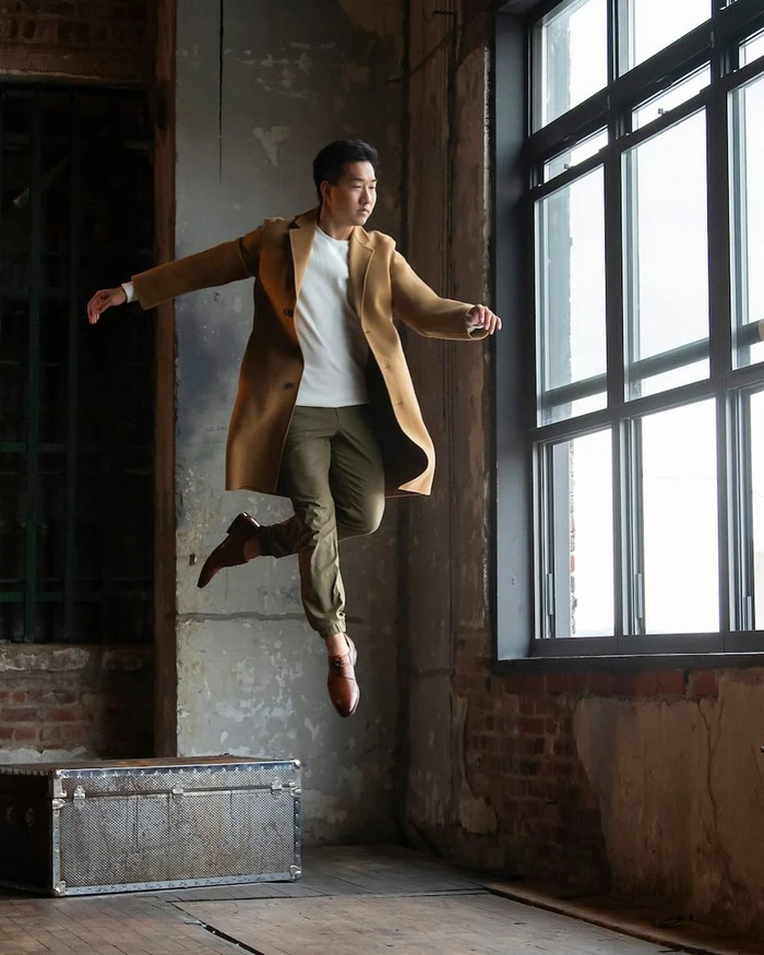 Man in camel coat leaping mid-air in front of tall industrial windows by Rachel Neville