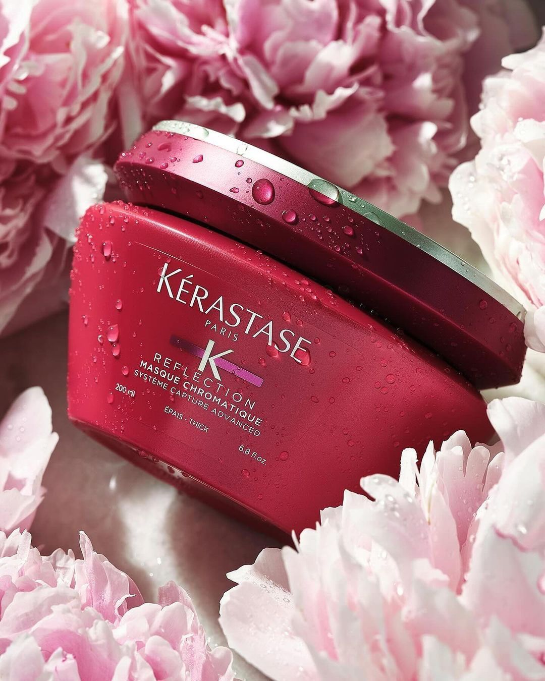Close-up product shot of Kérastase hair masque with red container and lid, surrounded by soft pink peony flowers and water droplets.