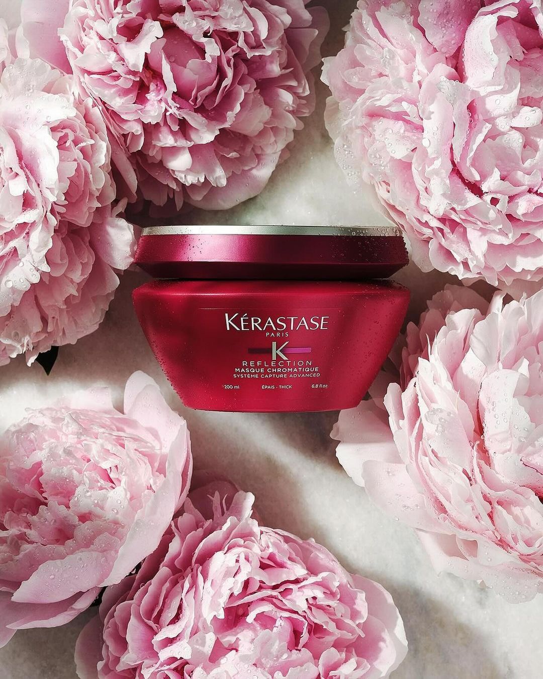 Kerastase hair product jar displayed in a composition with pink peony flowers on a neutral surface in a beauty and fashion shot.