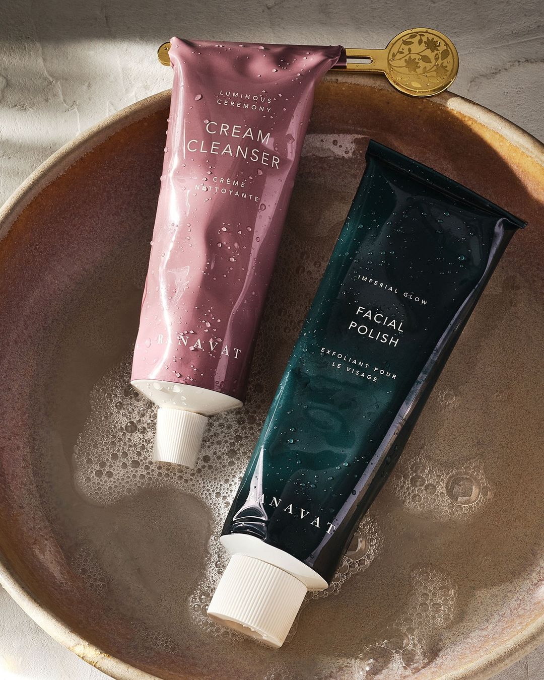 Two NAVAT skincare tubes, a cream cleanser and facial polish, sit in a beige ceramic bowl with bubbly water. Golden floral detail visible.