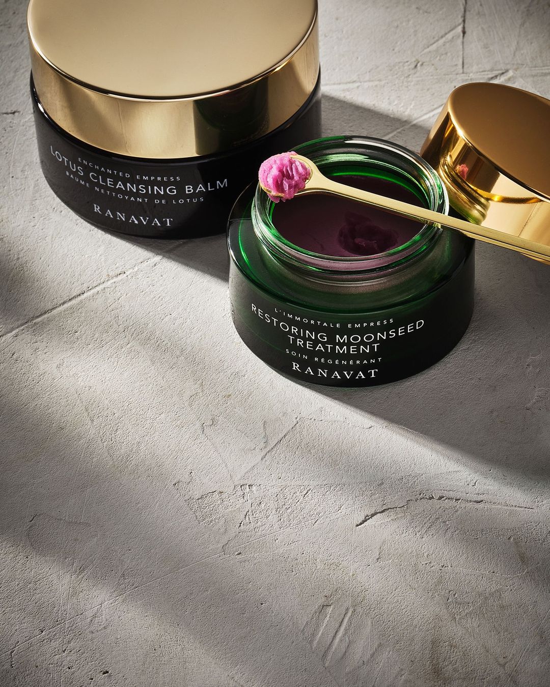 Cosmetic product placement featuring two jars of Ranavat skin cream, with golden lids, on a textured grey surface. Close-up shot.