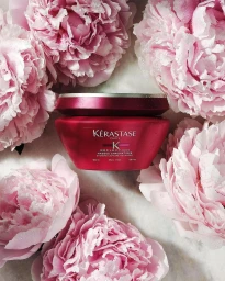 A deep pink Kerastase hair mask jar surrounded by fresh dewy pale pink peony flowers in a flat lay photograph.