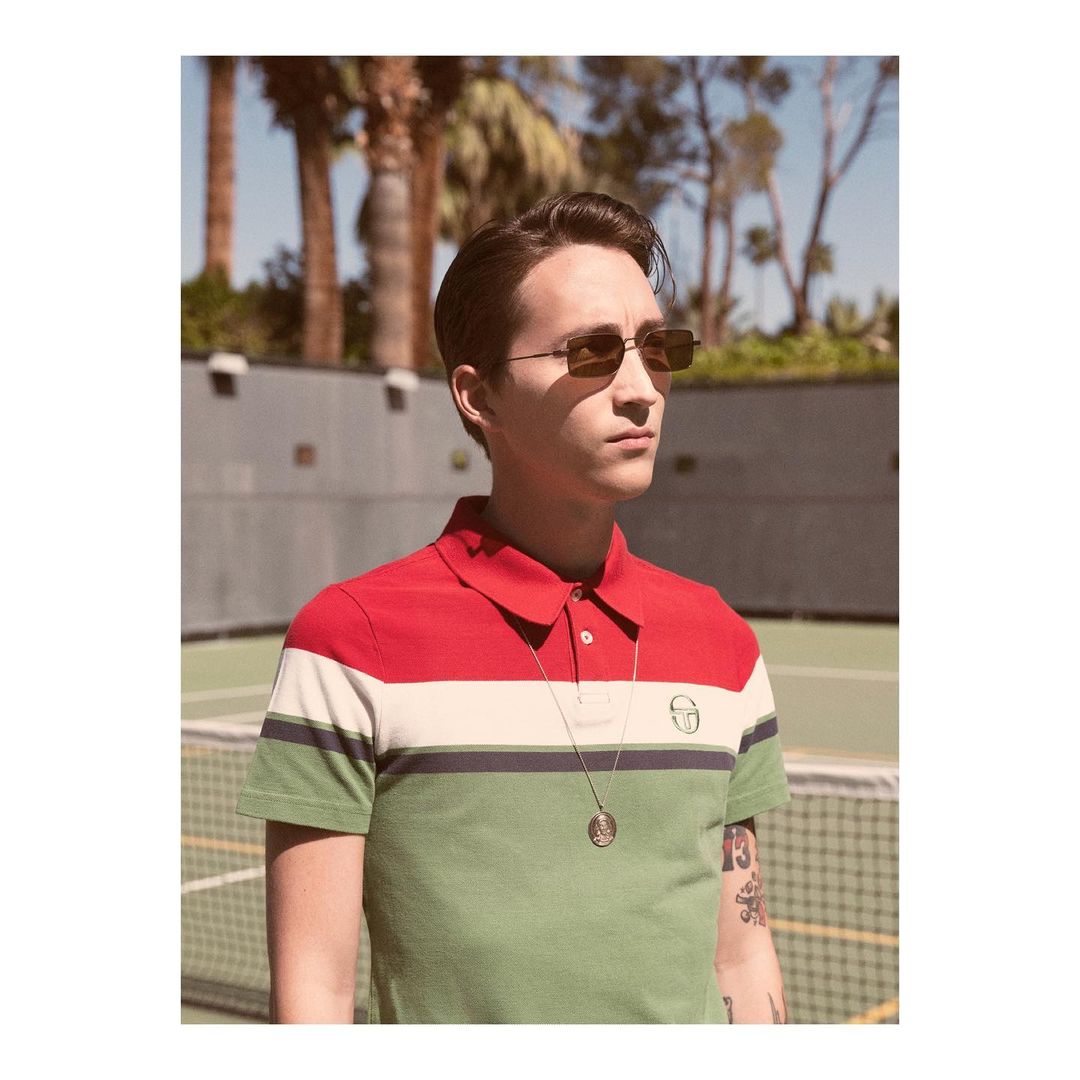 Portrait of a young adult male wearing sunglasses and a striped polo shirt, posing outdoors in front of a tennis court and trees.