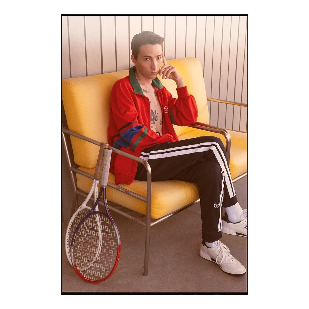Stylish young adult sits on a yellow chair with tennis rackets, wearing a red jacket and black and white striped pants indoors.