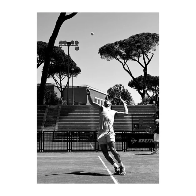 Black and white action shot of a tennis player serving on an outdoor court, with trees, stadium seating, and a tennis ball visible.