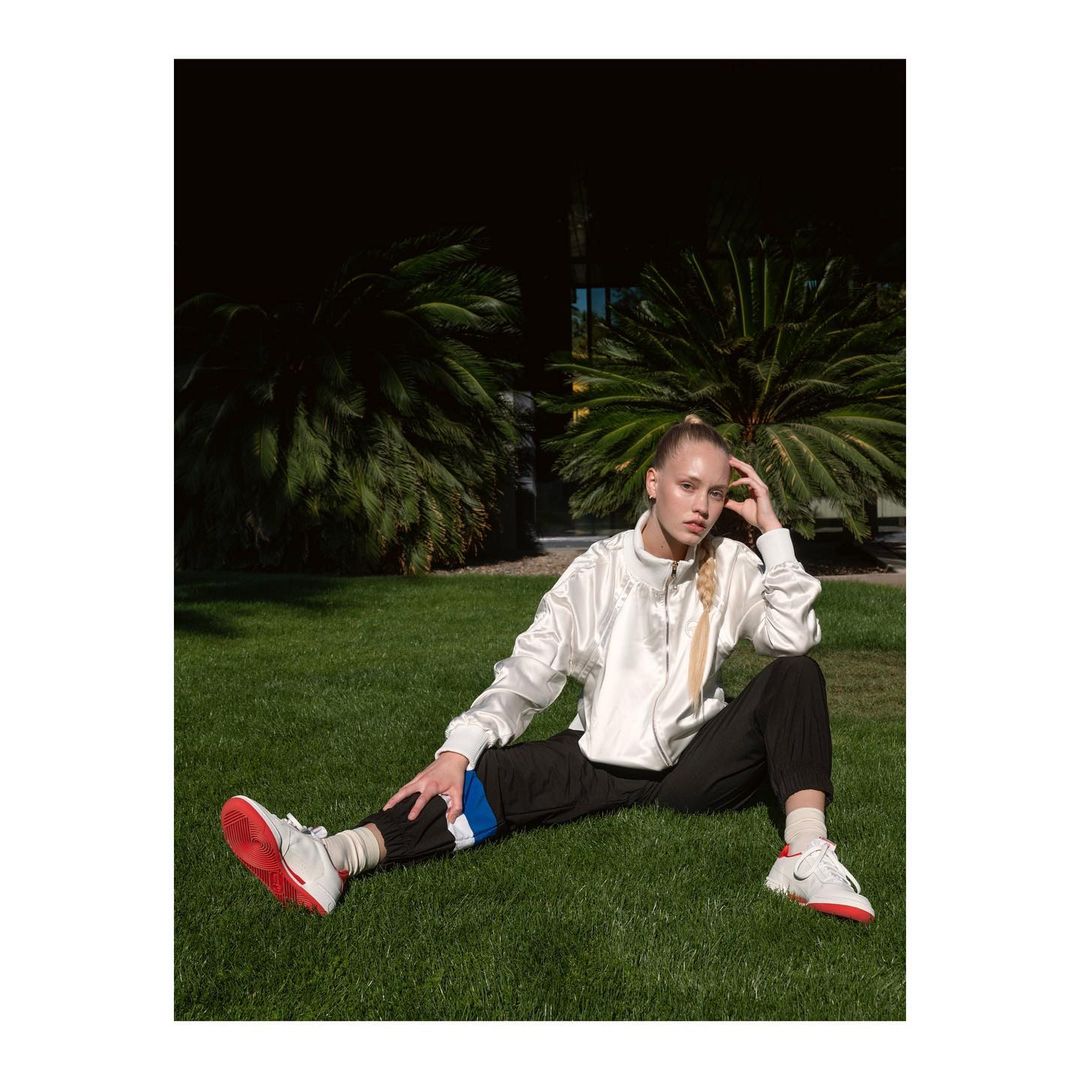 Woman in sportswear sitting on grass in front of palm trees. She wears white and red sneakers, black pants, and a white zip-up jacket.