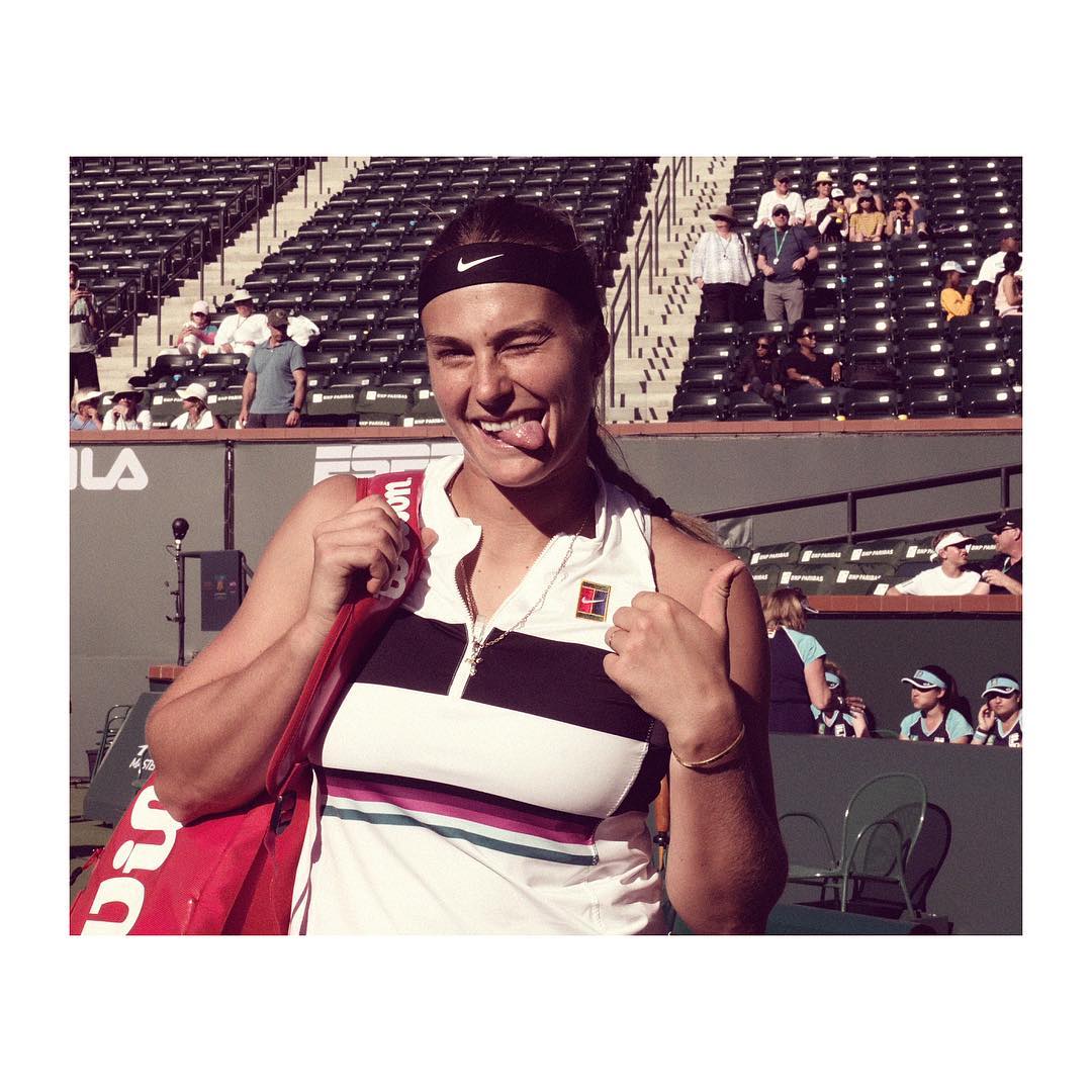 Female tennis player with tongue out after game at an outdoor tennis stadium on a sunny day, carrying a red tennis racquet bag.