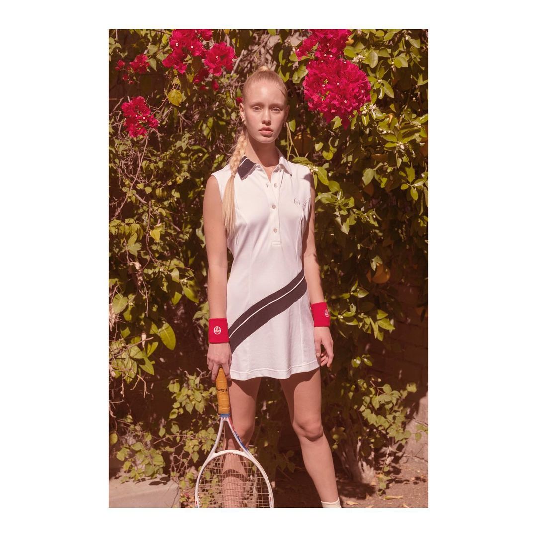 An adult woman stands outdoors in a white tennis dress, holding a racket, with bougainvillea in the background, creating a sports fashion photo.