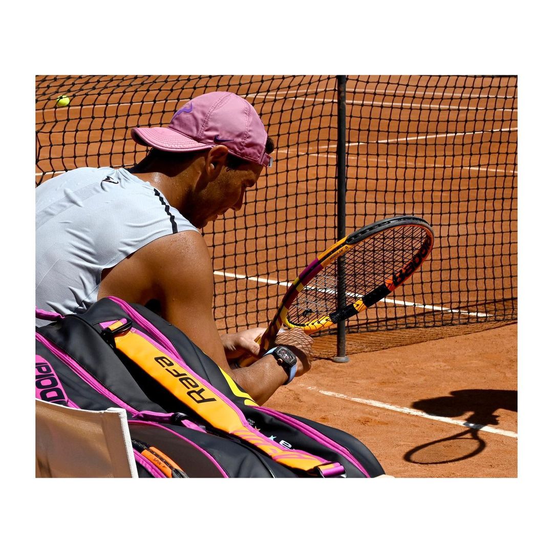 Tennis player with cap sits near net with racket and bag. Ground-level view on clay court with tennis ball in the distance.