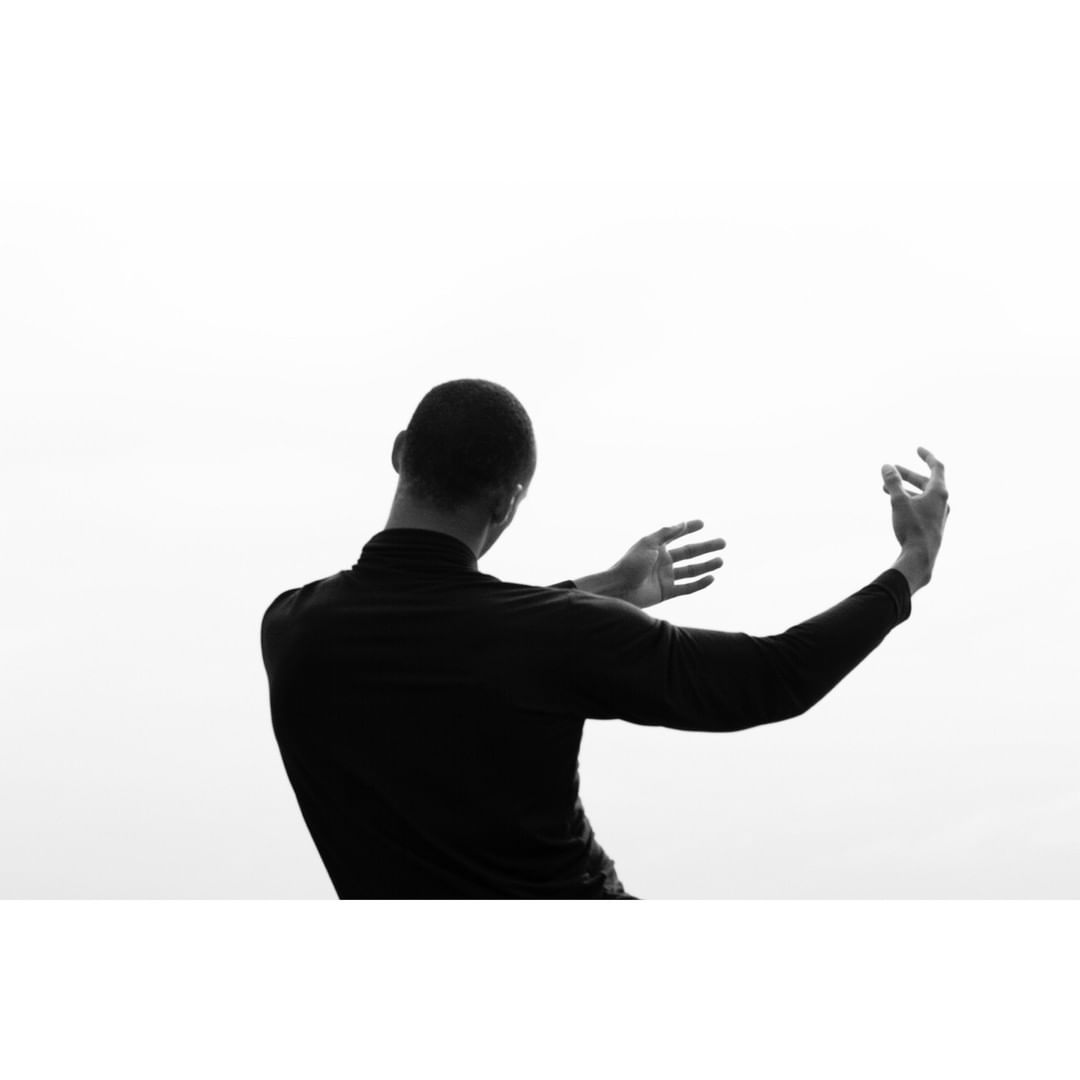 Back view of a Black adult male in dark clothing with arms raised against a bright, diffused background in a black and white photograph.