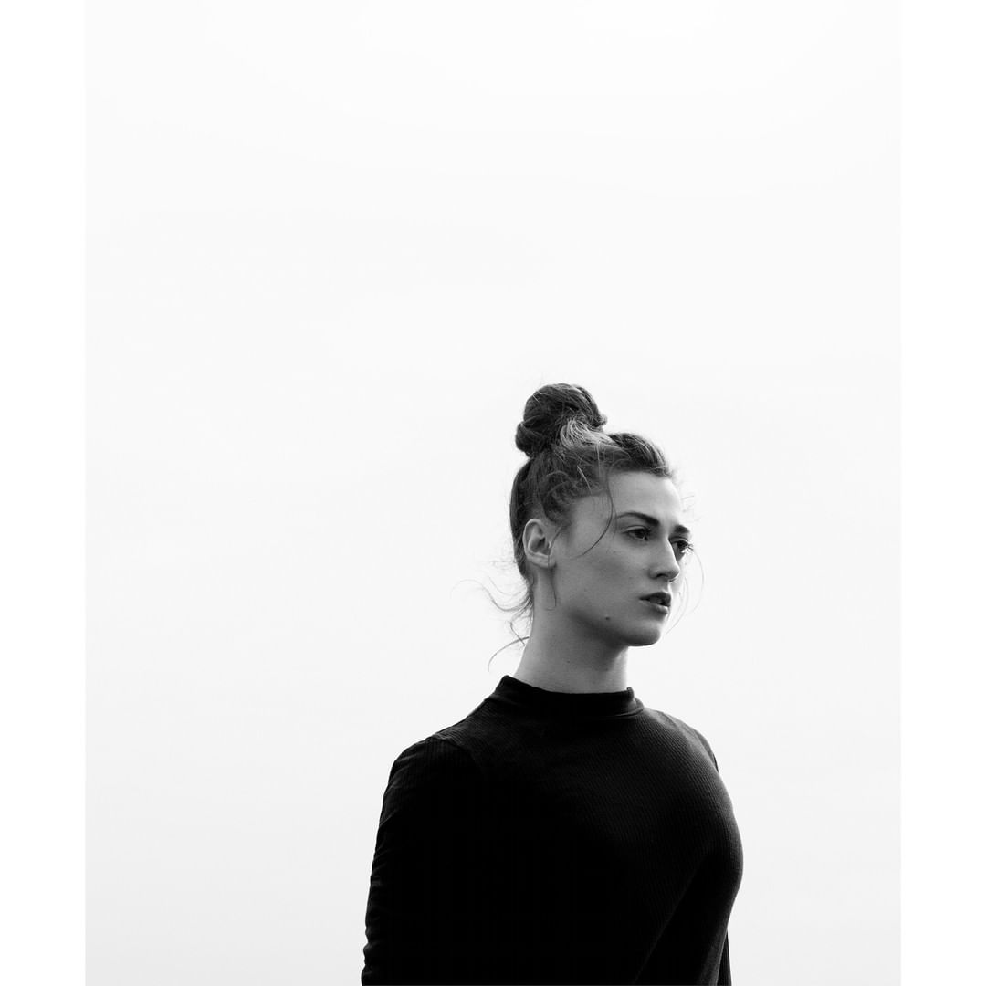 Portrait of a young woman with a bun hairstyle, wearing a dark top, in a black and white, minimalist outdoor setting against a bright sky.