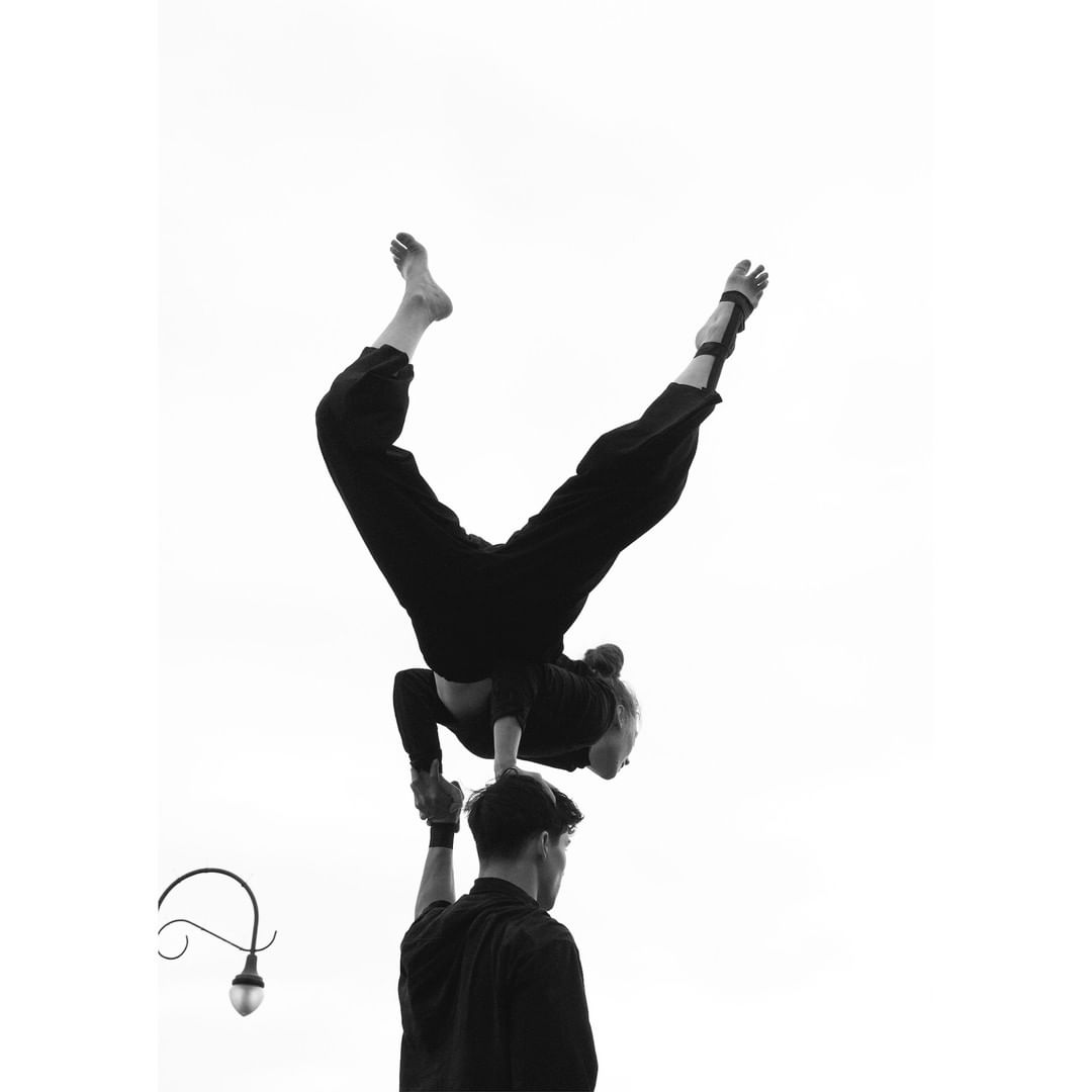 Two acrobats perform a handstand. A woman balances upside down on one hand of a man wearing matching black sportswear.