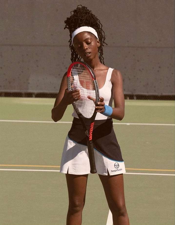 Young woman on green tennis court in white tennis dress holding red and black Wilson tennis racket, looking down. Outdoor, daytime.
