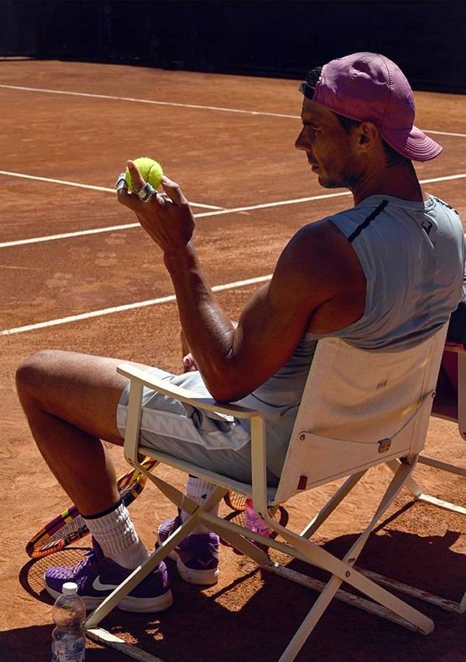 Professional tennis player Rafael Nadal resting on a chair on a clay court, holding a tennis ball, wearing a pink cap and purple tennis shoes.