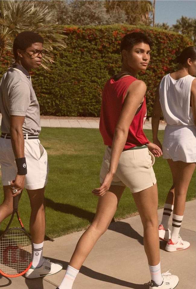 Three young adults in retro tennis outfits walk on a sidewalk lined with manicured grass and trees during a sunny day with filtered light.