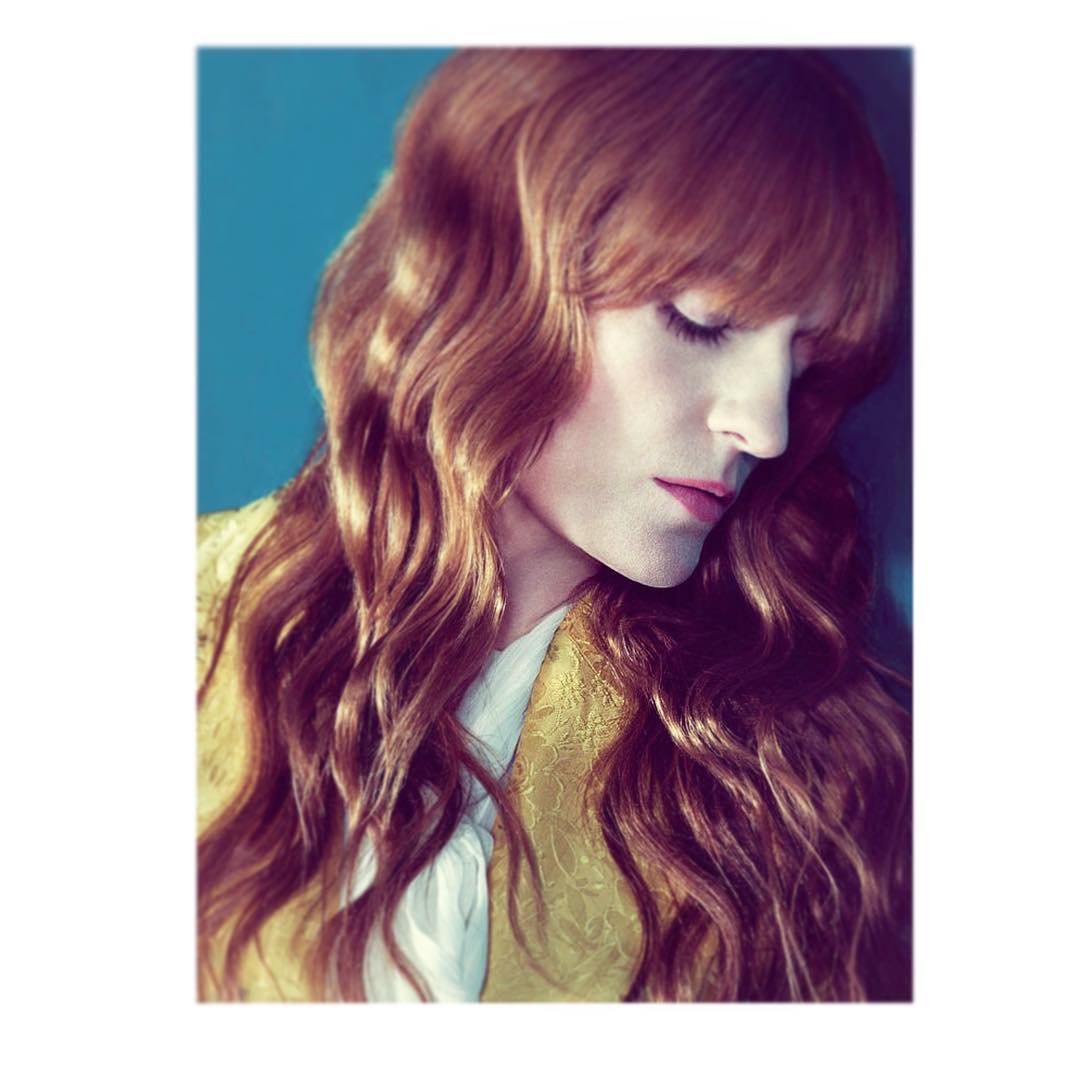 Portrait of a woman with wavy auburn hair, wearing a gold textured jacket, posing against a solid blue background in soft, diffused lighting.