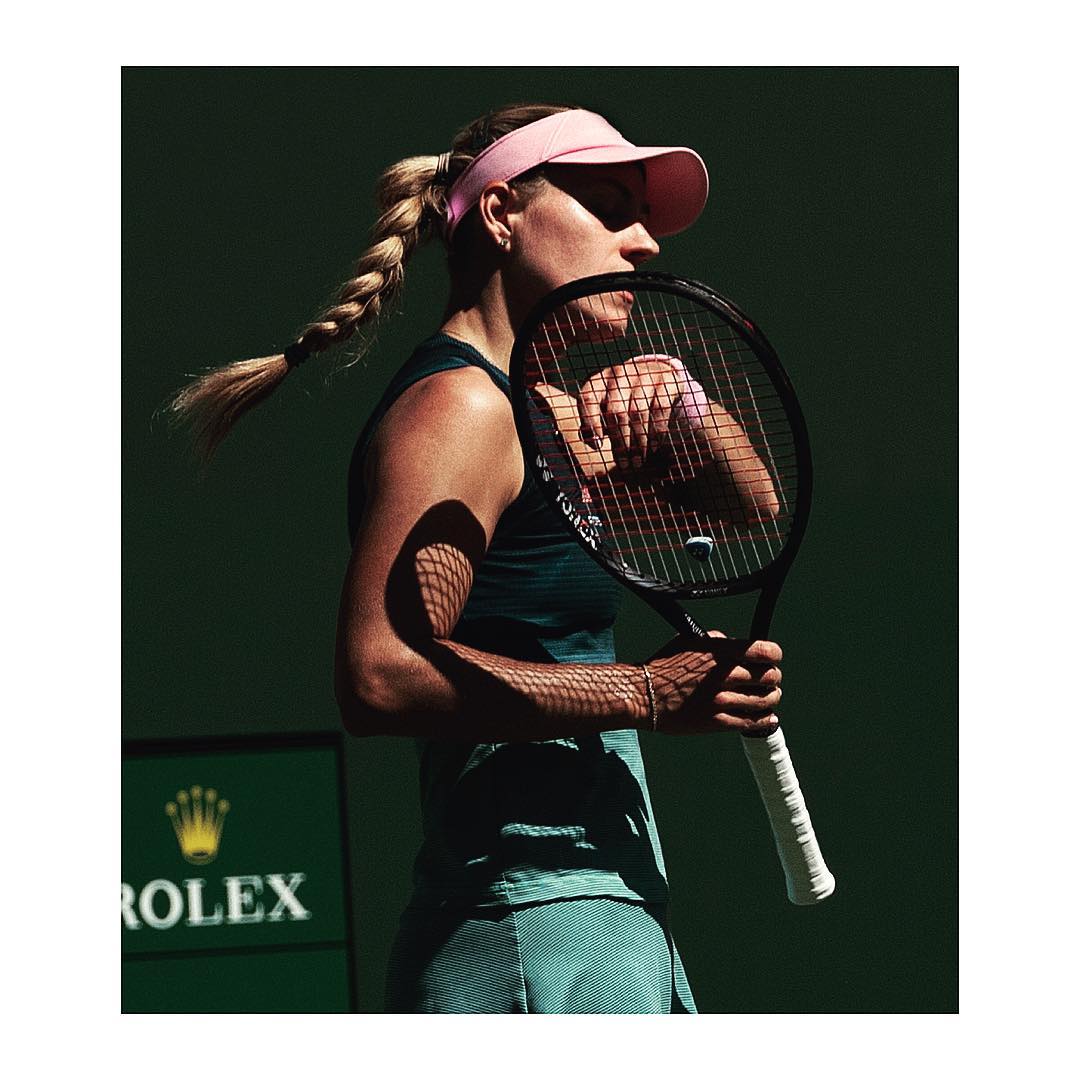 Female tennis player stands in a green court with a tennis racket and wearing a pink visor. Close up shot against a dark green background.