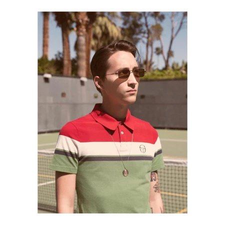 Man wearing sunglasses and a colour blocked polo shirt standing outdoors by a tennis court with palm trees