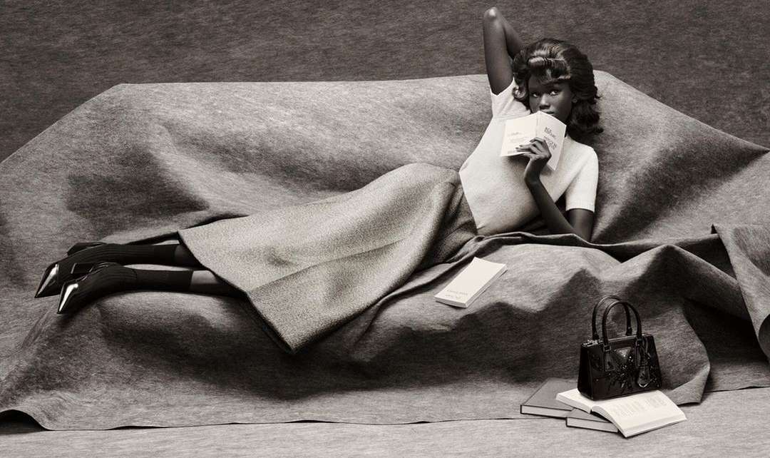 Elegant woman lounges on a draped couch in a studio wearing a skirt and t-shirt, reading with books and purse nearby.