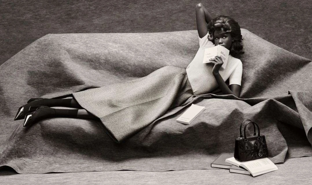 Elegant woman lounges on a draped couch in a studio wearing a skirt and t-shirt, reading with books and purse nearby.