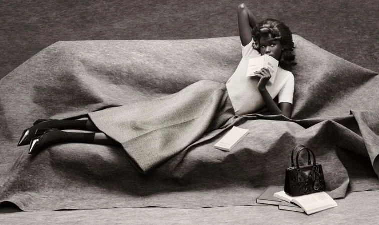 Elegant woman lounges on a draped couch in a studio wearing a skirt and t-shirt, reading with books and purse nearby.