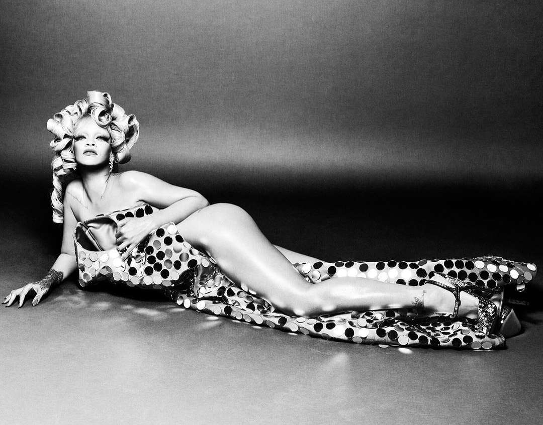 Monochrome portrait of a woman lying down in a polka dot dress and hair rollers, striking a pose against a gray gradient backdrop.