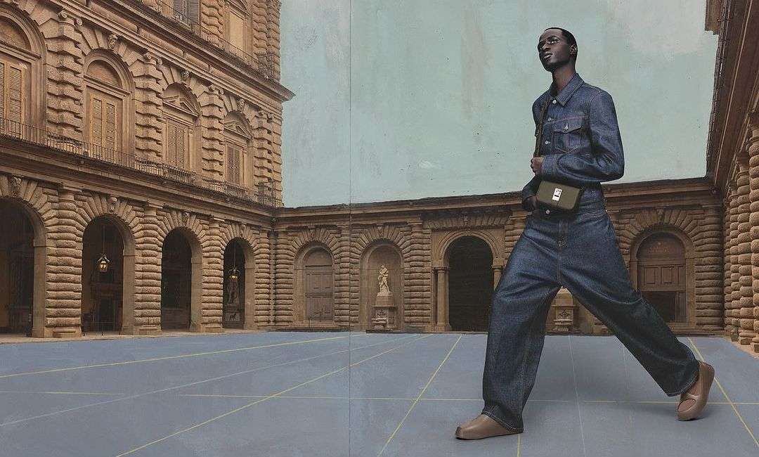 Fashion shot: A Black male model in full denim outfit poses with a green satchel bag in a courtyard with classic architecture in the background.