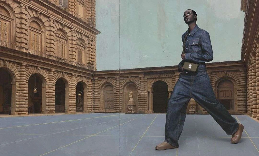 Fashion shot: A Black male model in full denim outfit poses with a green satchel bag in a courtyard with classic architecture in the background.