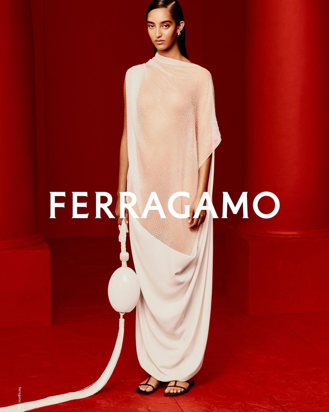 Fashion model wears a beige asymmetric dress with a white spherical bag, standing in a photo studio with a vibrant red backdrop.