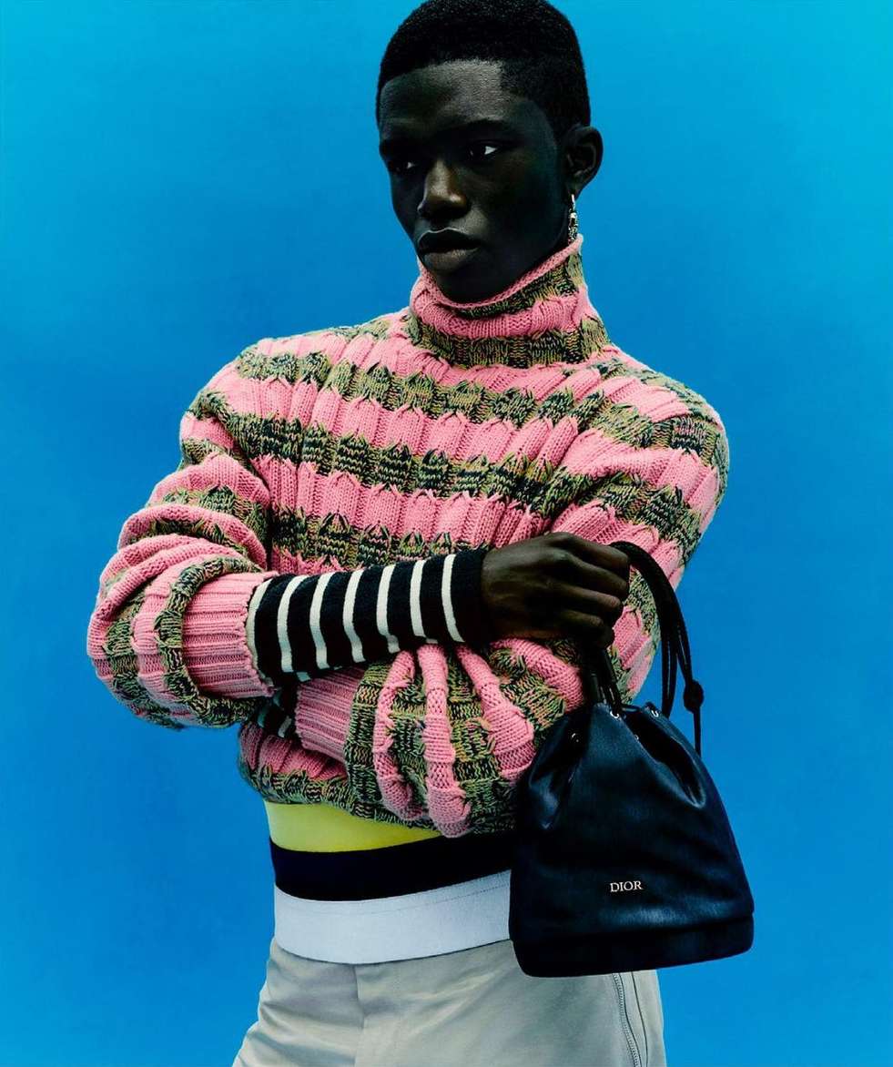 Studio shot of a dark-skinned adult male model wearing a pink striped sweater, light pants, and a black Dior purse against a blue background.