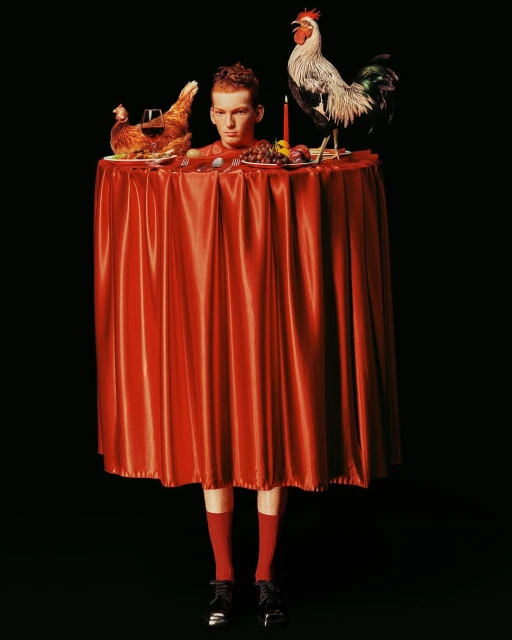 Surreal image of a red-haired person with a table draped in red fabric, featuring cooked and live chickens, in a dark studio.