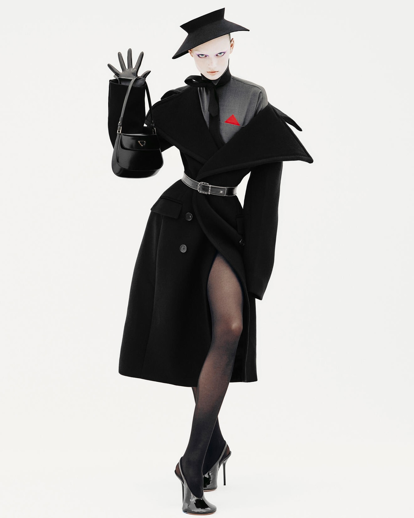 Fashion editorial shot of an adult female wearing a black coat, hat, gloves, and heels, posing with a black purse on a white background.